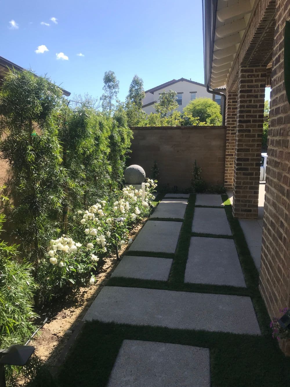 Pathway lined with white flowers and greenery beside a brick house under a clear blue sky.