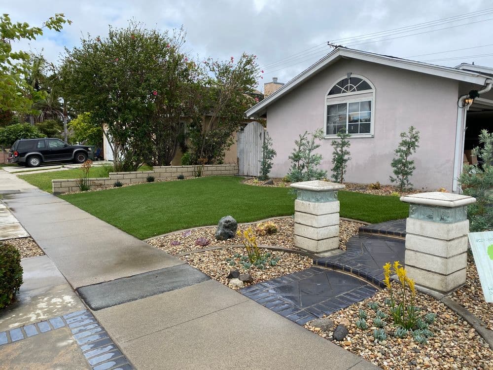 Residential front yard with artificial grass, stone planters, and landscaped gravel details.